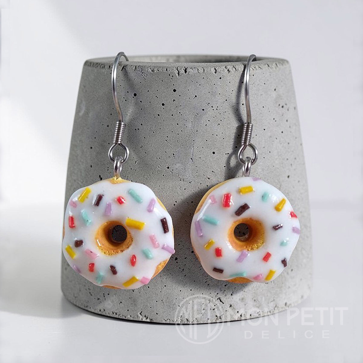 White donut earrings with colored sprinkles on a gray cement base. Miniature food jewelry.